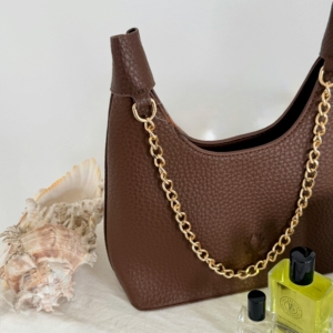 Stylish brown handbag with gold chain detail, paired with perfumes and seashells in a minimalist setting.