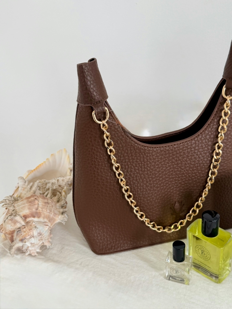 Stylish brown handbag with gold chain detail, paired with perfumes and seashells in a minimalist setting.